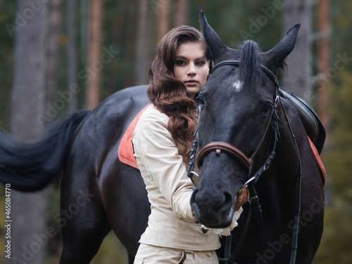 A young beautiful brunette rider next to a black horse in full ammunition, portrait in a stable in a forest area © Ulia Koltyrina