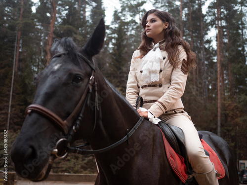 A young beautiful brunette rider in an elegant retro suit riding a black horse, in a forest area