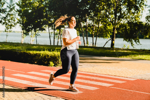 Full size portrait of a body positive plus-size plump woman athlete jogging running in fitness outfit, losing weight while listening to music in stadium outdoors