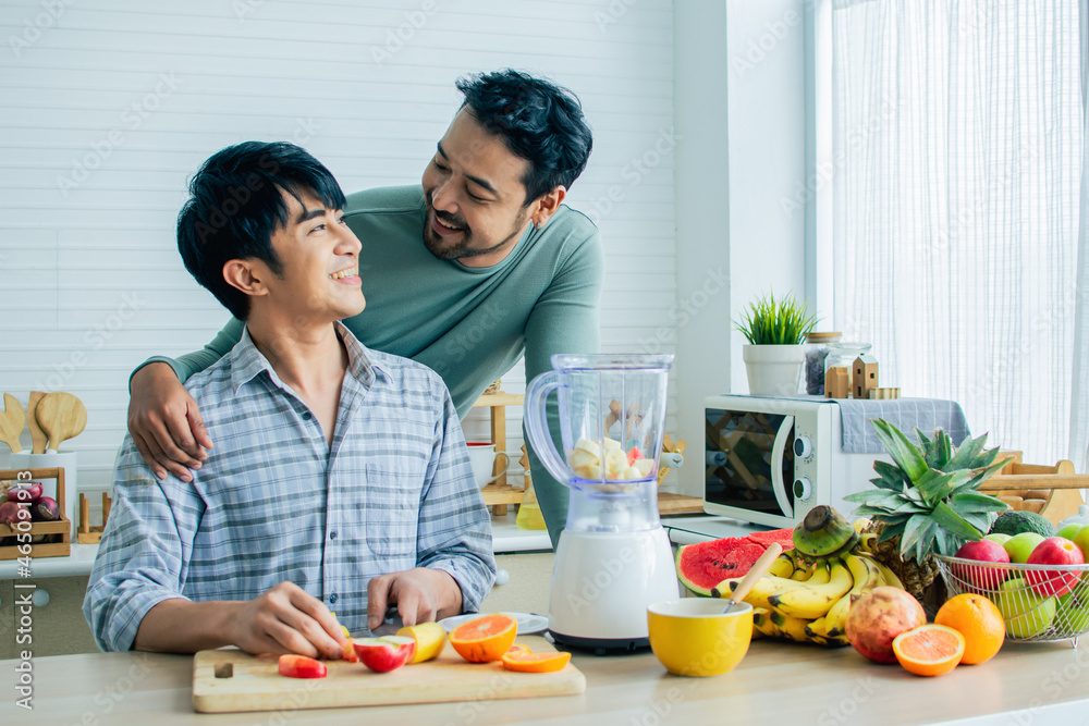Gay LGBT sweet Asian couple wearing pajamas, smiling, looking each other, hugging with happiness and love while making breakfast with healthy fruits in kitchen at home in morning. Lifestyle Concept.