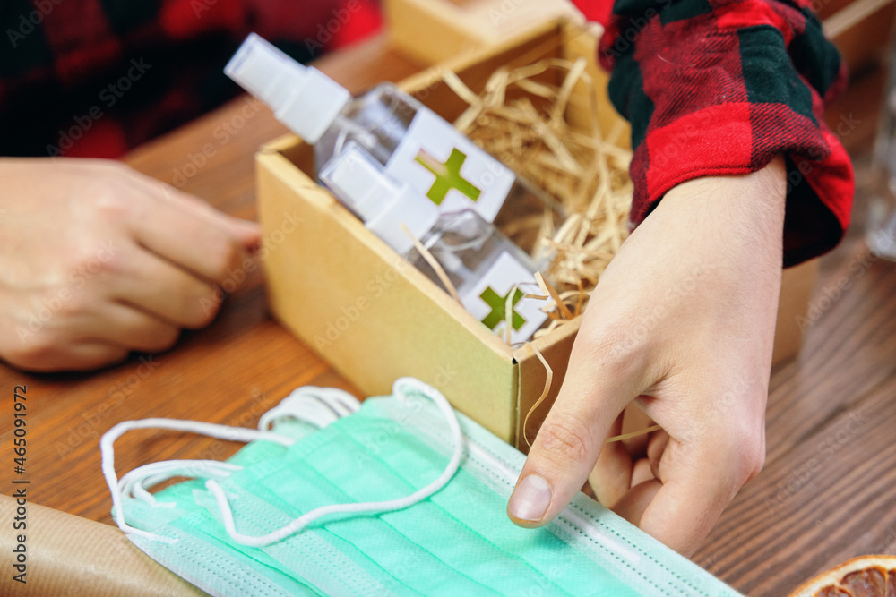Fototapeta premium Men's hands take medical masks to put them in a gift box with antiseptics. Gift in quarantine on a wooden table. Sticker with a green cross on the sanitizers.