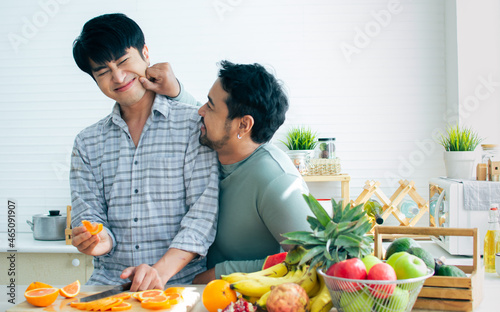 Gay LGBT sweet Asian couple wearing pajamas, smiling, teasing and pinching cheek with happiness and love, eating healthy fruits for breakfast in kitchen at home in the morning. Lifestyle Concept.