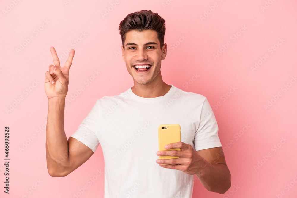 Young caucasian man holding a mobile phone isolated on pink background joyful and carefree showing a peace symbol with fingers. Young caucasian man holding a mobile phone isolated on pink background joyful and carefree showing a peace symbol with fingers.
