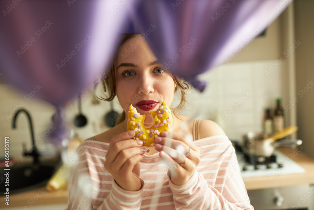 Selective focus of adorable charming female enjoying her doughnut looking at camera through violet helium balloons, isolated against comfortable modern kitchen. Favorite food, festive decoration
