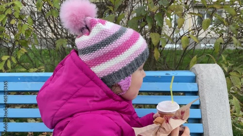 5 years old Girl sitting on blue bench and eating waffles and drinking juice. Autumn pink clothes. Eating outdoors.