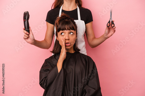 Young mixed race woman getting a haircut at the hairdresser isolated on pink background is saying a secret hot braking news and looking aside