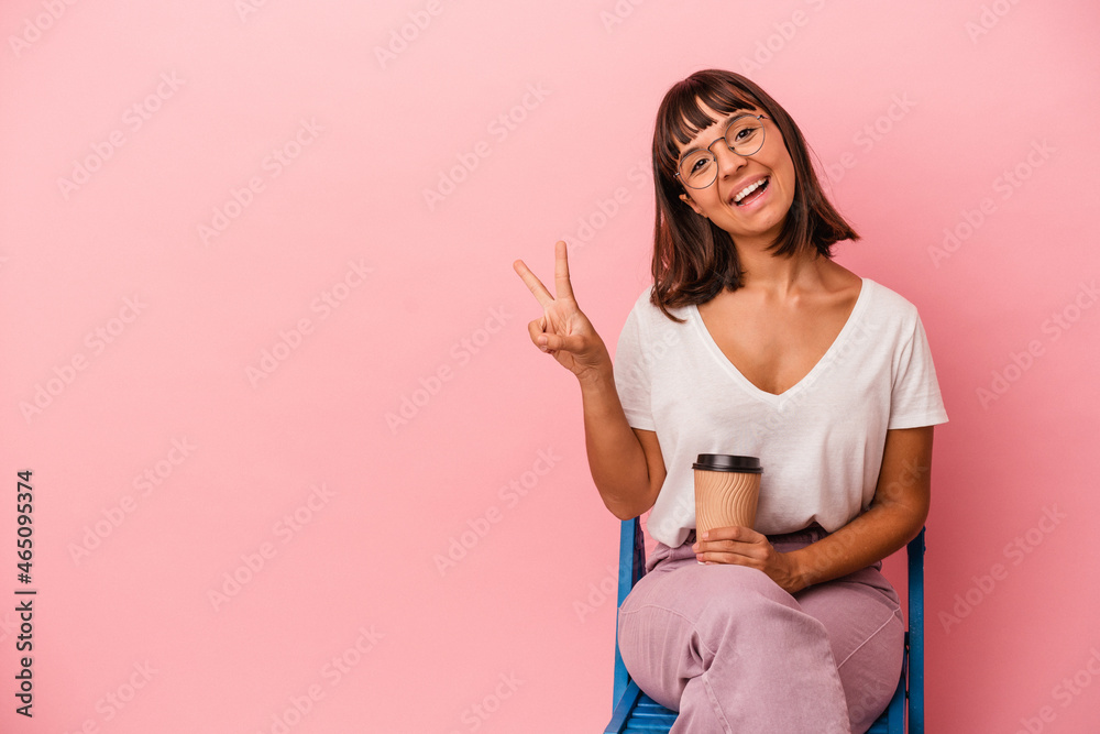 Young mixed race woman sitting on a chair holding a coffee isolated on pink background joyful and carefree showing a peace symbol with fingers. Young mixed race woman sitting on a chair holding a coffee isolated on pink background joyful and carefree showing a peace symbol with fingers.