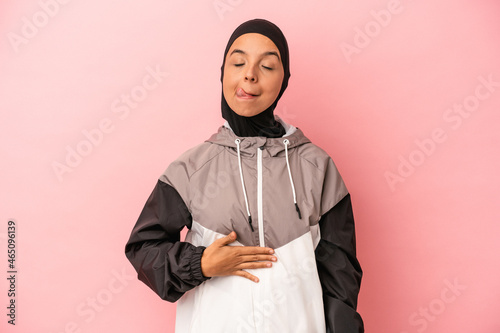 Young Arab woman with sport burqa isolated on pink background touches tummy, smiles gently, eating and satisfaction concept.