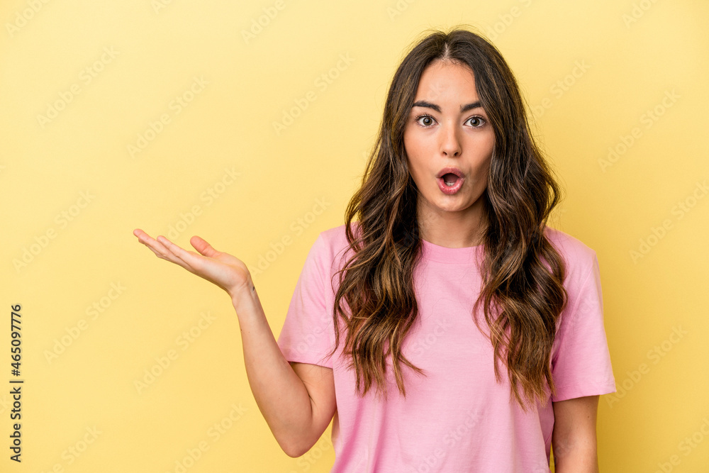 Young caucasian woman isolated on yellow background impressed holding copy space on palm. Young caucasian woman isolated on yellow background impressed holding copy space on palm.