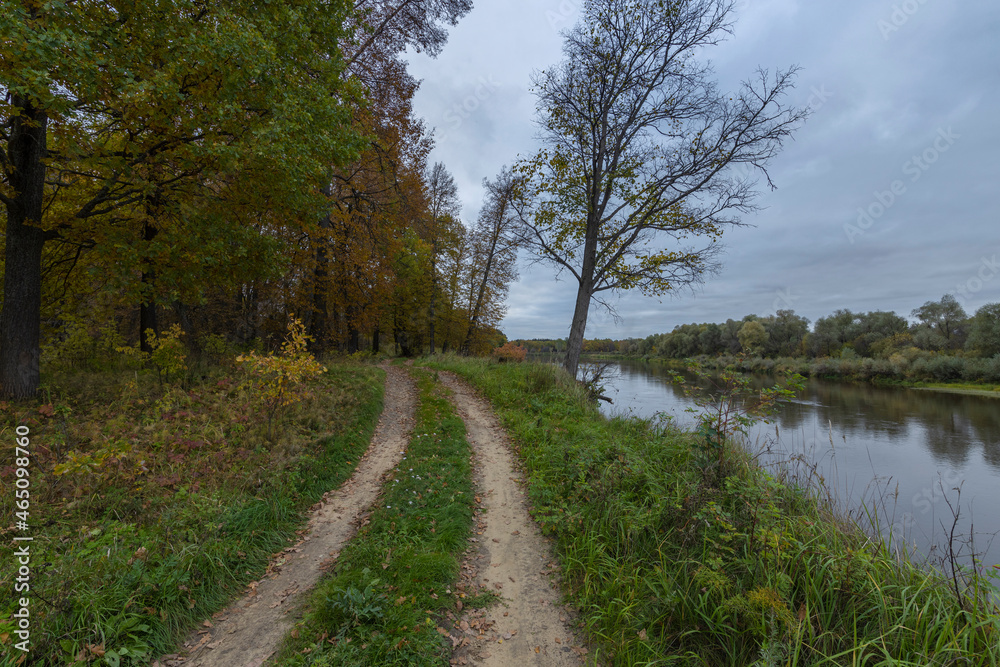 Dirt road along the river bank. Cloudy autumn weather. Rural landscape with road, river and autumn forest.