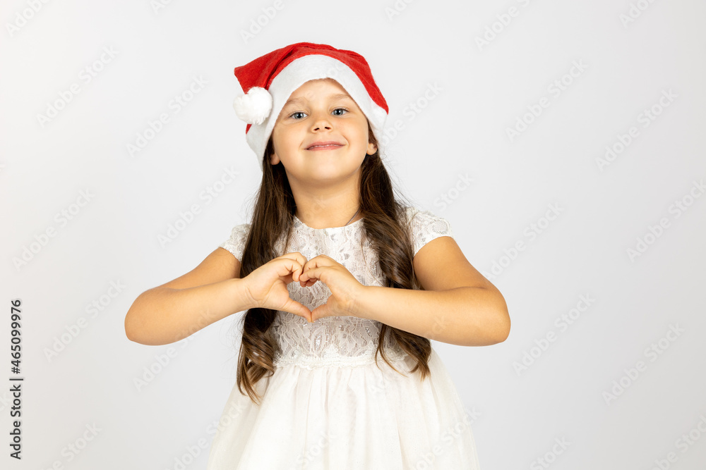 portrait of smiling, beautiful girl in white dress, wearing red Santa Claus hat shows heart sign from fingers isolated on white background. portrait of smiling, beautiful girl in white dress, wearing red Santa Claus hat shows heart sign from fingers isolated on white background.