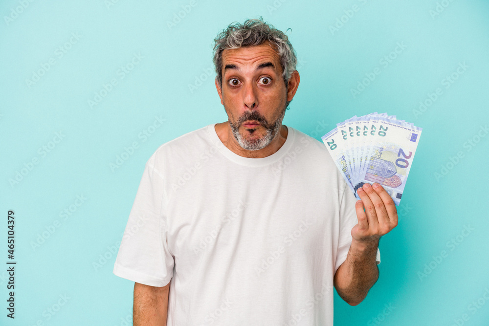 Middle age caucasian man holding bills isolated on blue background shrugs shoulders and open eyes confused. Middle age caucasian man holding bills isolated on blue background shrugs shoulders and open eyes confused.