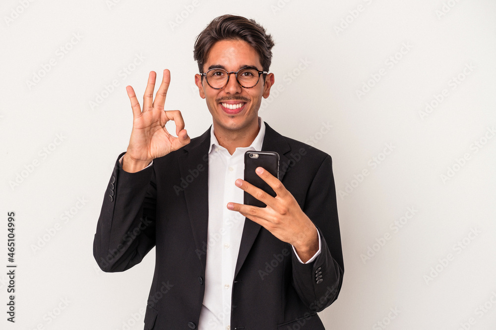 Young mixed race business man holding mobile phone isolated on white background cheerful and confident showing ok gesture. Young mixed race business man holding mobile phone isolated on white background cheerful and confident showing ok gesture.