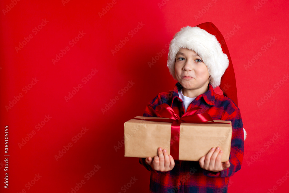 Happy holiday and Merry Christmas. Portrait of a boy in a cap with gifts in his hands on a red background. A place for text. Happy holiday and Merry Christmas. Portrait of a boy in a cap with gifts in his hands on a red background. A place for text.