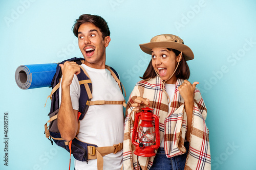 Young mixed race hiker couple isolated on blue background points with thumb finger away, laughing and carefree.
