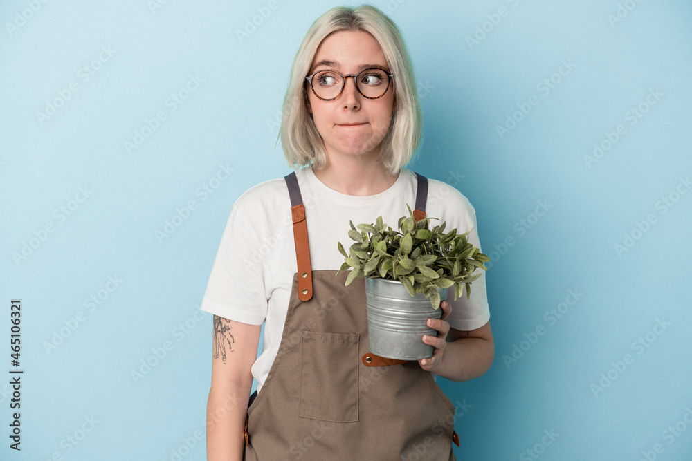 Young gardener caucasian woman holding a plant isolated on blue background confused, feels doubtful and unsure. Young gardener caucasian woman holding a plant isolated on blue background confused, feels doubtful and unsure.