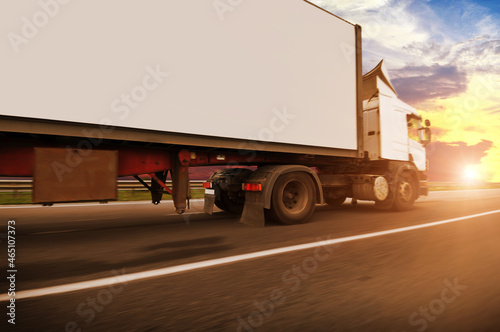Big truck with a white trailer driving fast on a countryside road against a sky with a sunset