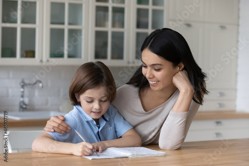 Happy mom giving help and support to schoolboy son while kid doing homework, learning writing exercise, training handwriting in home kitchen. Loving mother encouraging an hugging studying child