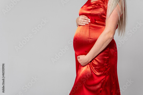Young pregnant woman, dressed in a red dress, holding hands on her belly on a white background. No face.