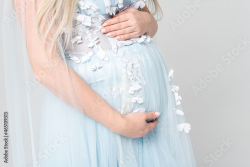 Young pregnant woman wearing a light blue dress holding hands on her belly on a white background. Without a face.