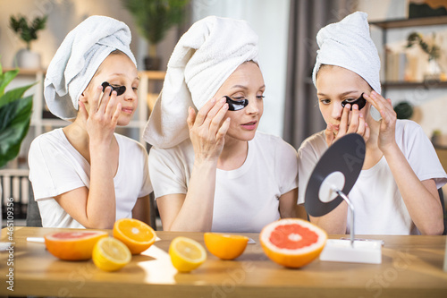 Two teen sisters sitting with their charming mother at table and applying black collagen pads under eyes. Moisturising procedures for face skin. Domestic treatment.