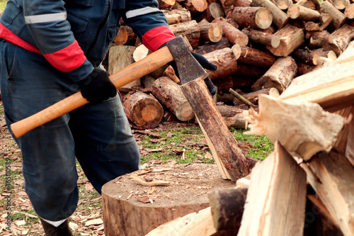 The concept of the onset of cold weather and fuel harvesting. An axe in the hands of a man, hands in work gloves, against the background of chopped firewood.