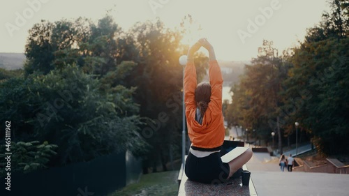 The tired girl stretches after a long period of online work. Dressed in a bright orange blouse, hair caught in a bun, positioned on the park strip. 4k concept