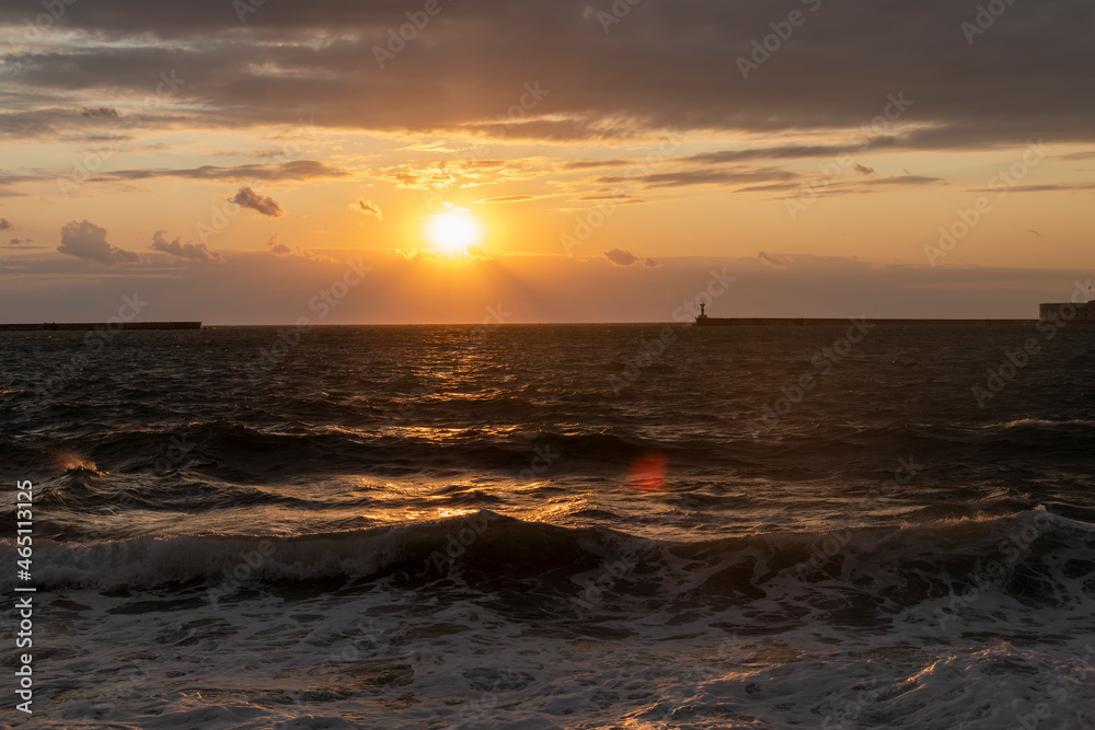 Orange sunset sky with few clouds above the stormy waves of the Black Sea tending to the shore. Silhouettes of two breakwaters and a lighthouse are visible on the horizon. Beauty in nature theme.