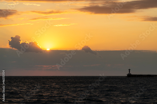 Orange sunset sky with few clouds above dark waves of the Black Sea tending to the shore. Silhouettes of breakwater with lighthouse are visible on the horizon. Beauty in nature theme.