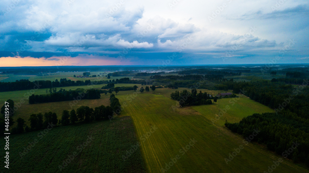 Aerial view of the sunset over green fields and forest. Heavy clouds in the sky. 