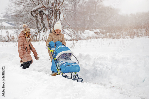 young woman with baby stroller and her daughter walk in the park in snowfall in winter