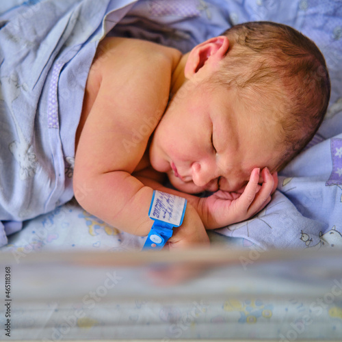 A newborn baby with a maternity hospital bracelet on his arm is sleeping in a crib. A newly born child in a clinic bed behind a transparent glass photo