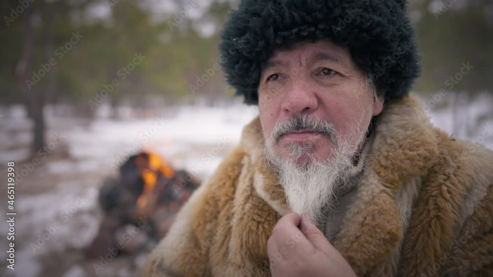 Old confident indigenous man stroking grey beard looking away smiling. Portrait of senior man sitting in winter forest with fire burning in slow motion at background. Lifestyle and freedom Old confident indigenous man stroking grey beard looking away smiling. Portrait of senior man sitting in winter forest with fire burning in slow motion at background. Lifestyle and freedom