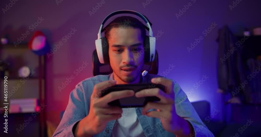 Happy asia man gamer wear headphone competition play video game online with smartphone colorful neon light in living room at night modern house. Esport streaming game online, Home quarantine activity.
