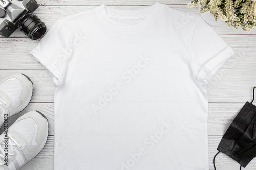 White womens cotton t-shirt mockup with sneaker, retro camera and face mask on white wooden background. Design t shirt template, print presentation mock up. Top view flat lay.