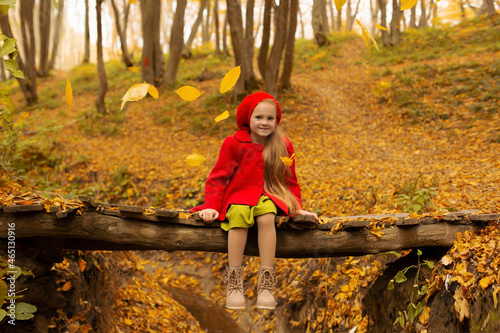 a beautiful happy little blonde girl in a red coat and a red beret is sitting on a bridge across a river in the city's autumn yellow park