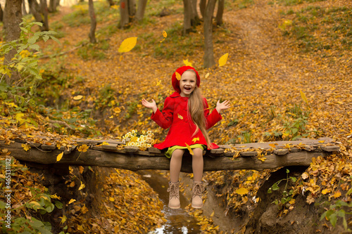 a beautiful happy little blonde girl in a red coat and a red beret is sitting on a bridge across a river in the city's autumn yellow park