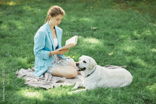 Full length portrait of young woman reading book outdoors with Labrador dog lying on grass in park, copy space