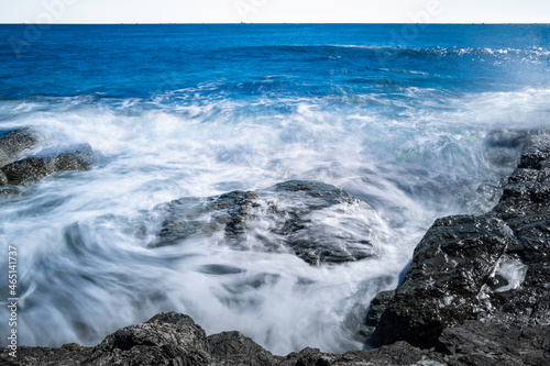 Smooth silky waves flow over the black rocks on the beach. Panoramic motion blur rocky shore image.