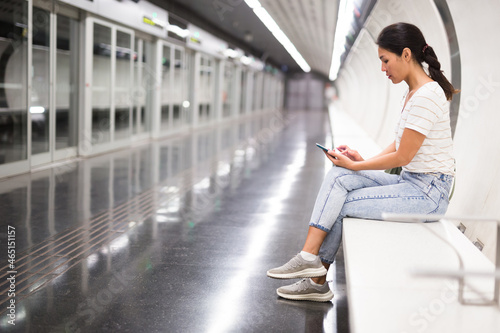 Asian woman with smartphone sitting on bench in subway station.