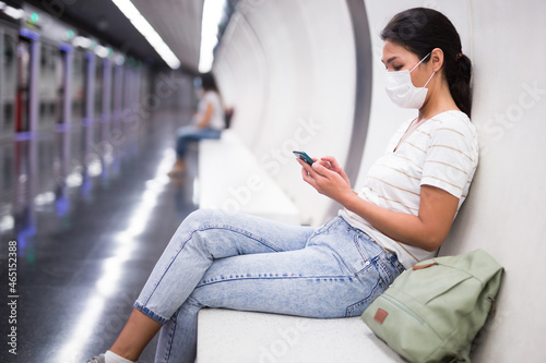 Asian woman in face mask with smartphone sitting in subway station and waiting for train.