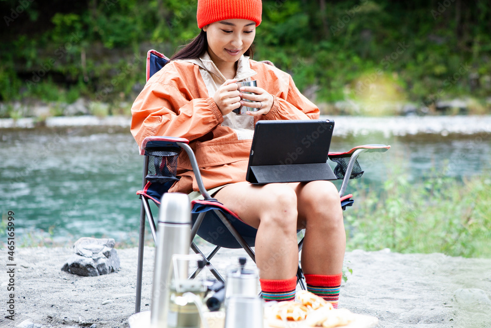 川辺でソロキャンプをする女性／デジタルタブレットで動画を見ている