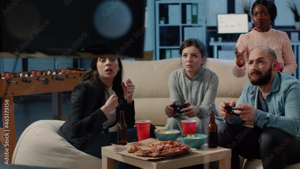 Coworkers playing video games with joysticks and losing after work at office. Colleagues enjoying game with controllers on television console for entertainment and drinks. People having fun Coworkers playing video games with joysticks and losing after work at office. Colleagues enjoying game with controllers on television console for entertainment and drinks. People having fun