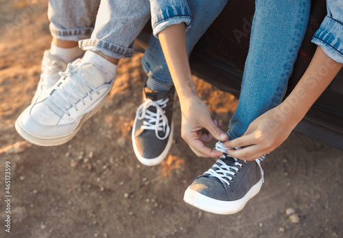 Close-up photo of legs in sneakers. The teenager boy and girl ties the laces on the sports shoes. Young people in casual blue denim clothes are sitting together outdoor.