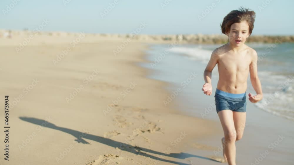 Long shot of boy in swimming trunks rolling ball along coastline. Tracking shot of happy and concentrated kid playing ball, running and exercising during holiday. Sport, summer vacation concept Long shot of boy in swimming trunks rolling ball along coastline. Tracking shot of happy and concentrated kid playing ball, running and exercising during holiday. Sport, summer vacation concept