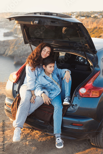 A young mother and her teenage son in denim enjoy the view of the sea and mountains on the top of the hill, sitting in the open trunk of a car and hugging together. Happy family relationship concept. © Evgeniya Grande