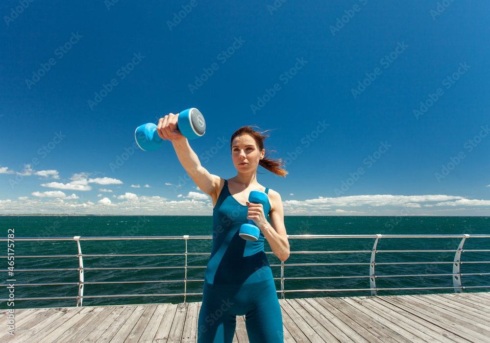 Sporty Caucasian woman exercising with dumbbells on the embankment at bright sunny day with blue sky and clouds. Healthy lifestyle concept. Wide angle Sporty Caucasian woman exercising with dumbbells on the embankment at bright sunny day with blue sky and clouds. Healthy lifestyle concept. Wide angle
