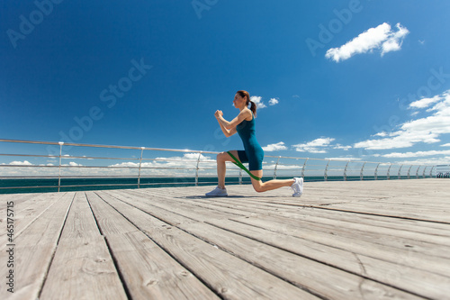 Fit woman doing exercise with fitness elastic bands, training legs on the embankment at bright sunny day with blue sky and clouds. Wide angle