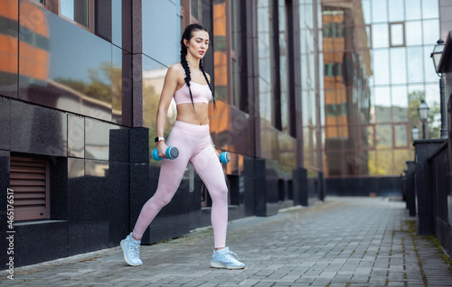 Sexy fit brunette woman with pigtails trains muscles with dumbbells in her hands on the background of a city building