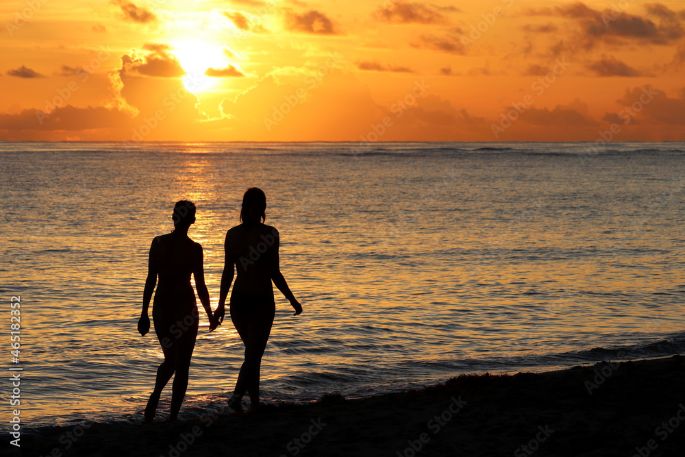 Silhouettes of two girls in bikini walking by the sand on evening ocean beach. Sunset on a coast, travel and holiday concept